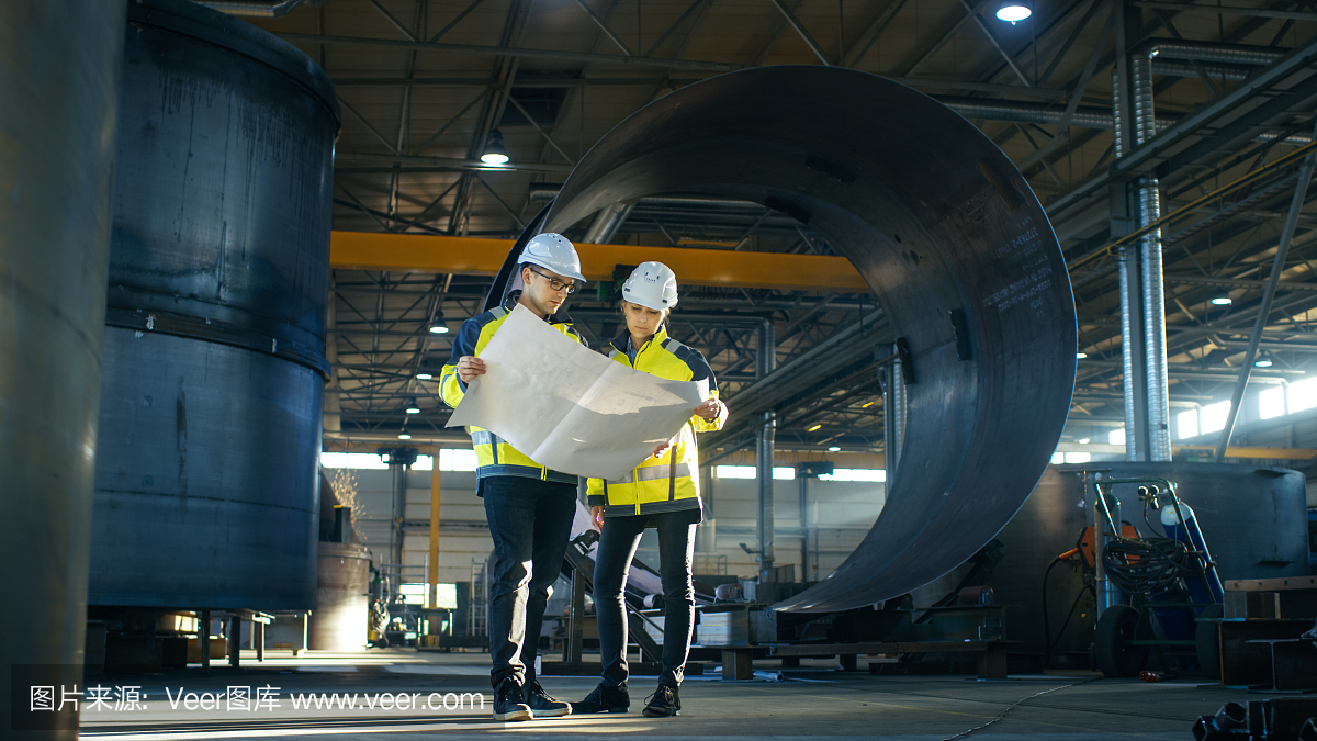 男和女工业工程师看着项目蓝图,而站在巨大的重工业制造工厂中间的管道部件